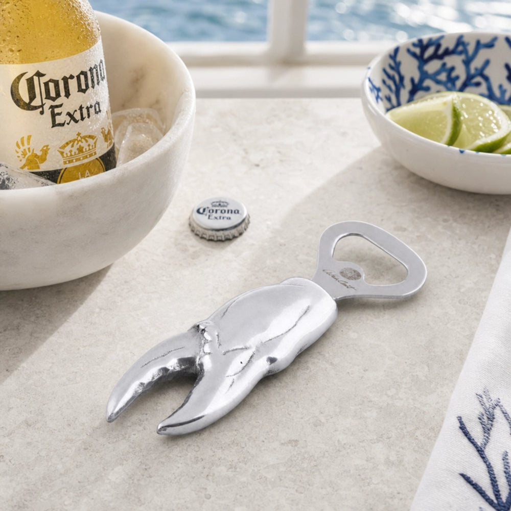 A silver claw-shaped bottle opener styled on a light stone surface, with a Corona Extra bottle cap nearby, a bowl of lime slices, and a bottle in a bowl in the background.