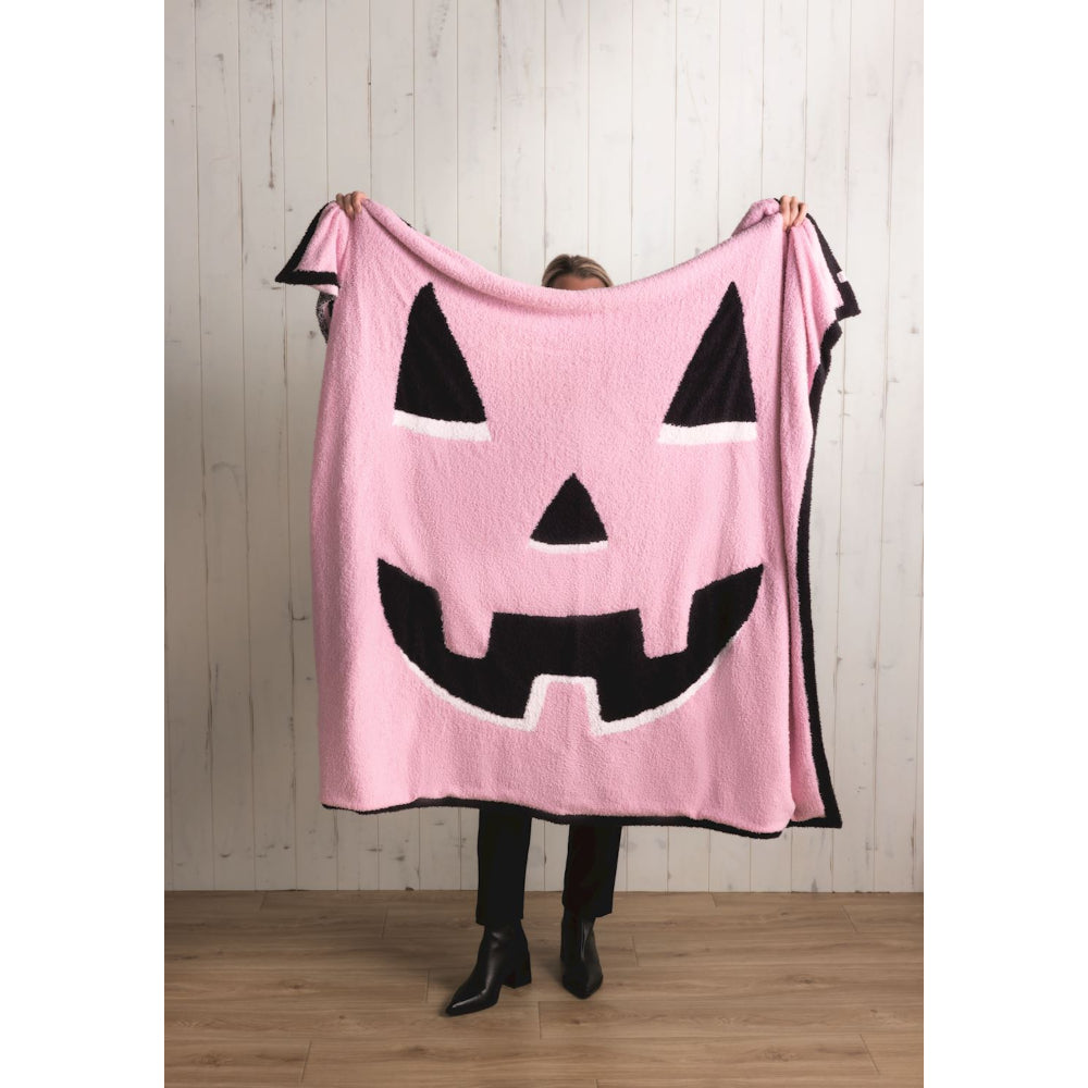 Person holding a pink blanket with a black jack-o'-lantern face design against a wooden wall.