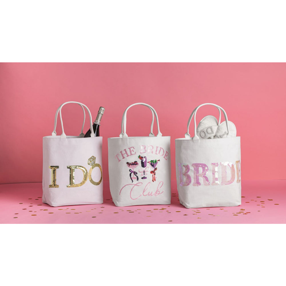 Three white fabric tote bags with white handles, decorated with pink, gold, and multicolor glitter text, displayed on a pink surface against a pink background.