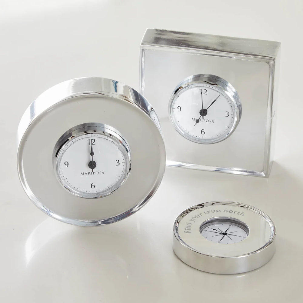 A set of three polished silver-toned metal desk accessories on a light cream background: a round clock with a white face and black hands, a square clock with a white face and black hands, and a circular compass with a white face and silver needle.