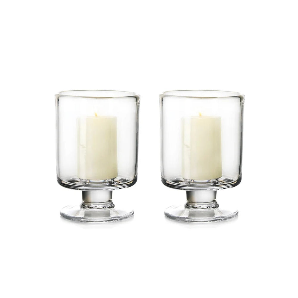 Two clear glass candle holders with pedestal bases, each containing a white pillar candle, shown on a plain white background.