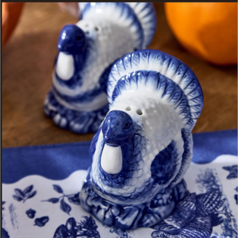 Blue and white ceramic salt and pepper shakers shaped like turkeys, displayed on a blue patterned cloth with a wooden surface and orange fruit in the background.