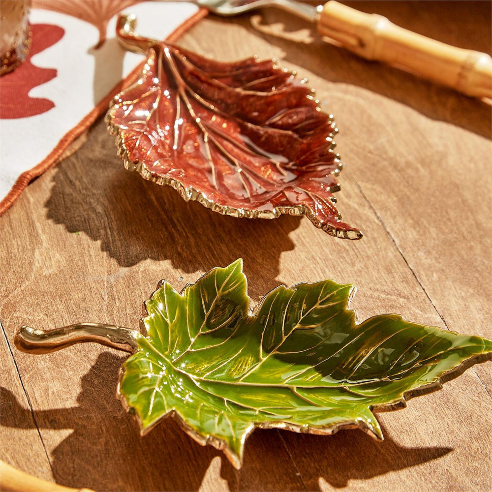Two decorative leaf-shaped serving dishes with metallic gold edges, one glossy green maple leaf and one glossy reddish-brown leaf, arranged on a wooden surface.