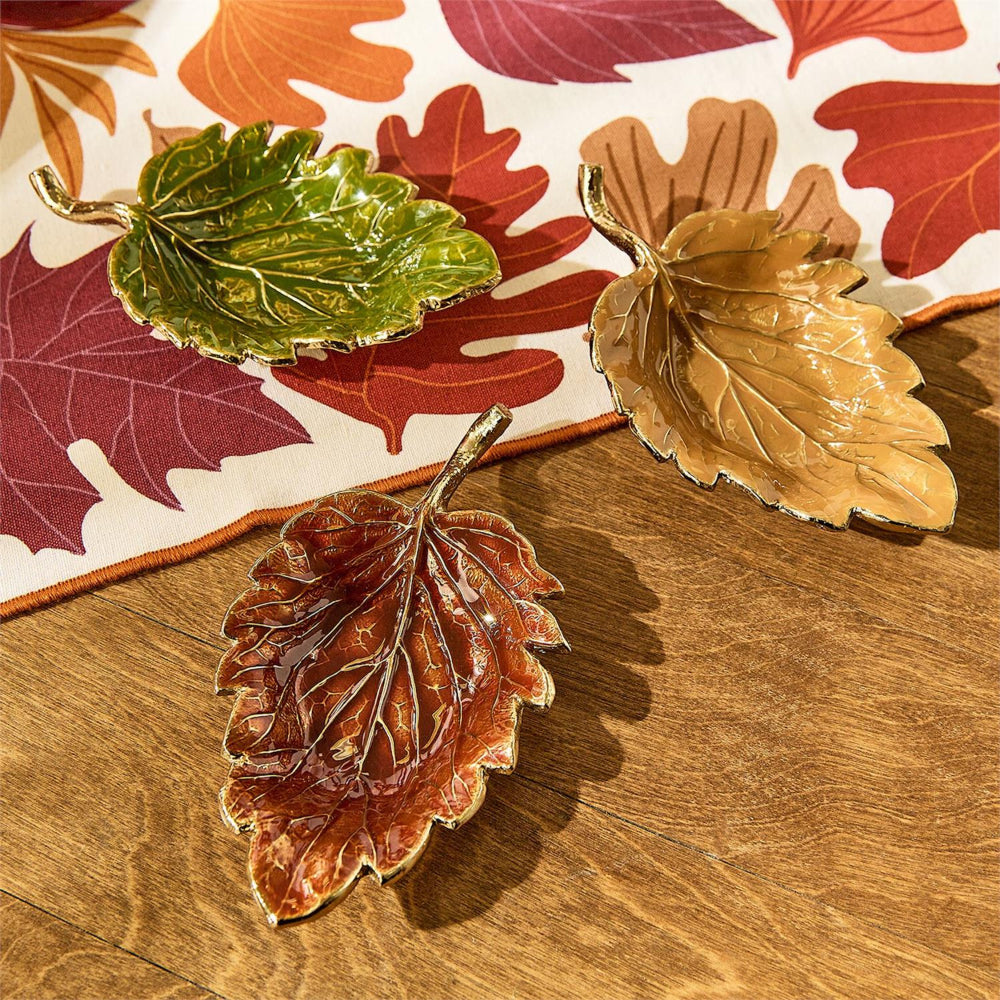 Three decorative leaf-shaped trays in green, tan, and reddish-brown with metallic gold edges, arranged on a wood floor over a patterned autumn leaf fabric.