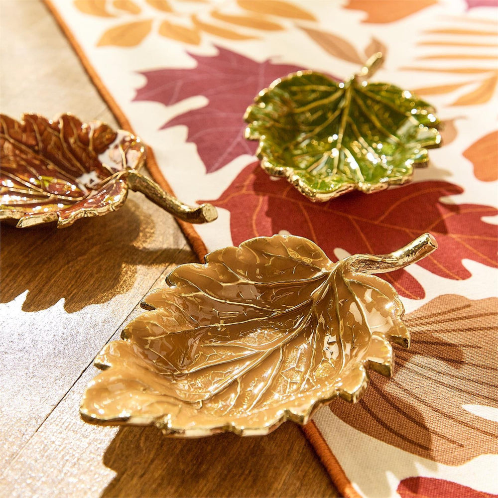Decorative leaf-shaped dishes in gold, green, and red on a patterned fabric background.