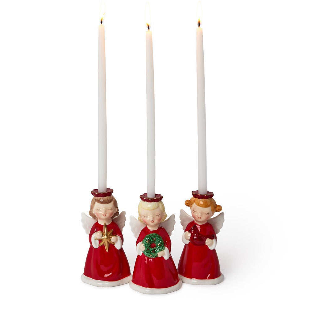 Three angel figurine candle holders in red dresses with white wings and red holos holding christmas decorations with long white candles on a white background.
