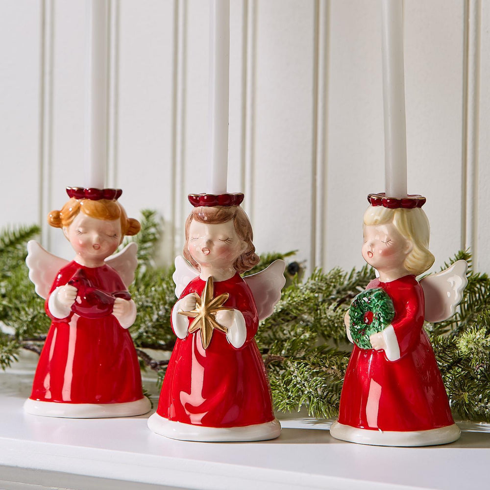 Three angel figurine candle holders in red dresses with white wings and red holos with long white candles holding a star, cardinal and wreath on a white surface with a decorative background.