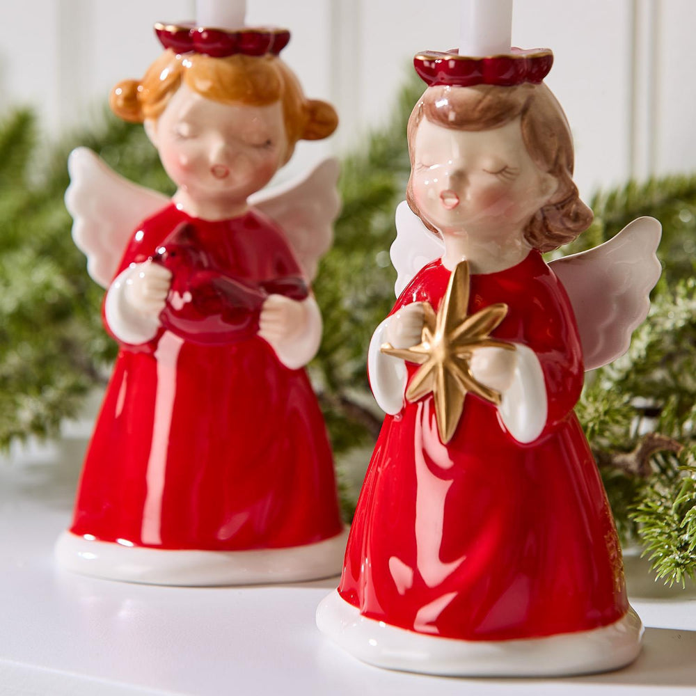 Two ceramic angel figurine candle holders in red dresses with white wings, one holding a star and the other a red cardinal, against a blurred greenery background.