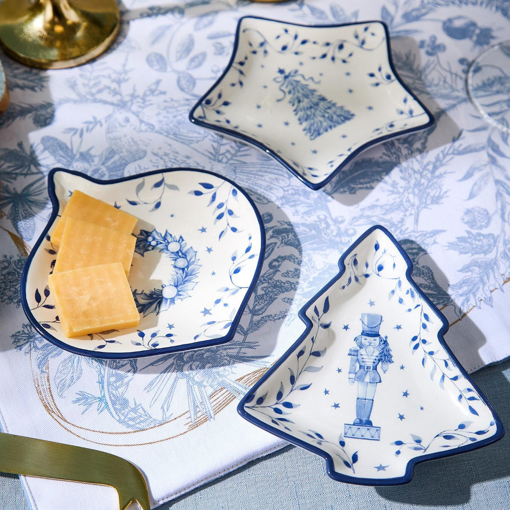 Three decorative christmas ceramic dishes with blue tree,wreath and nutcreaker designs on a blue and white tablecloth. 