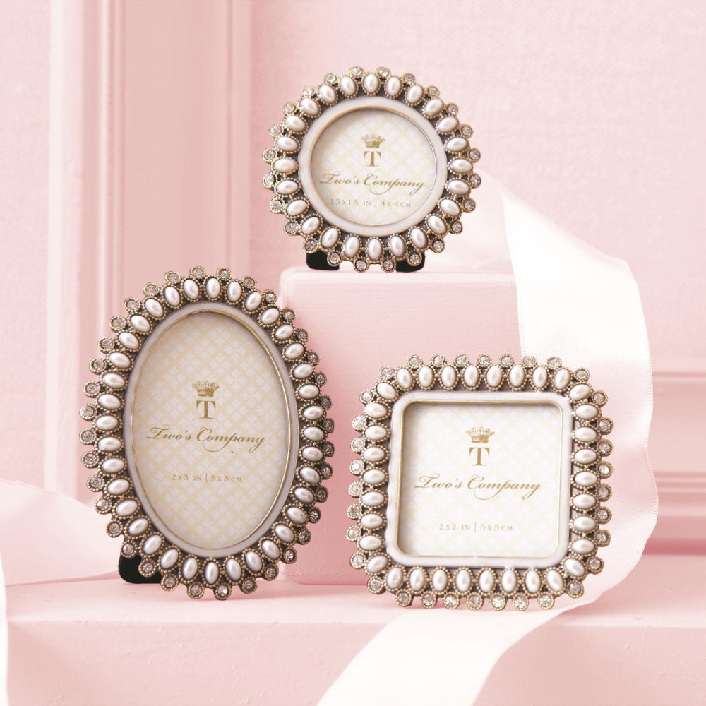 Three decorative picture frames with ornate silver-tone borders and clear rhinestones, displayed on a soft pink set. The oval frame stands on a small black stand, the round frame sits on a white pedestal, and the square frame rests on a pink ribbon.
