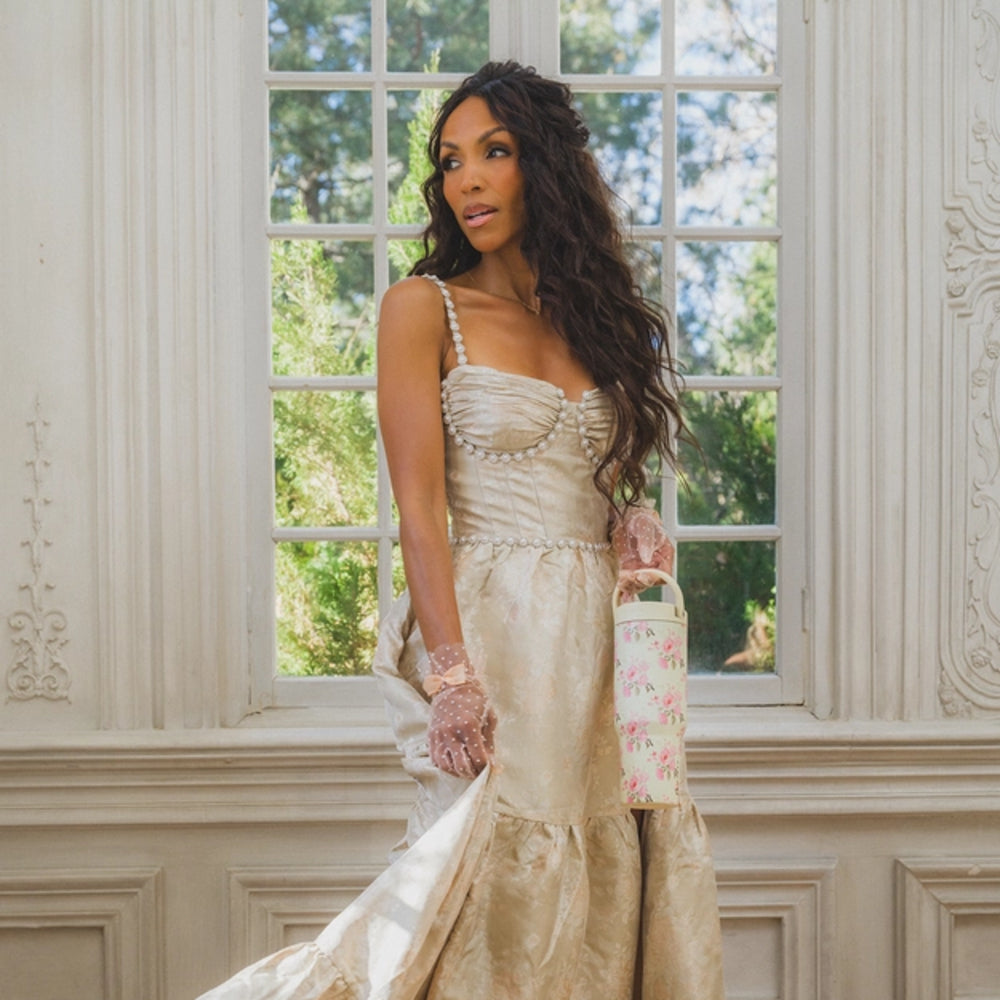 Woman in a beige dress holding a floral handbag in front of a window with greenery outside.