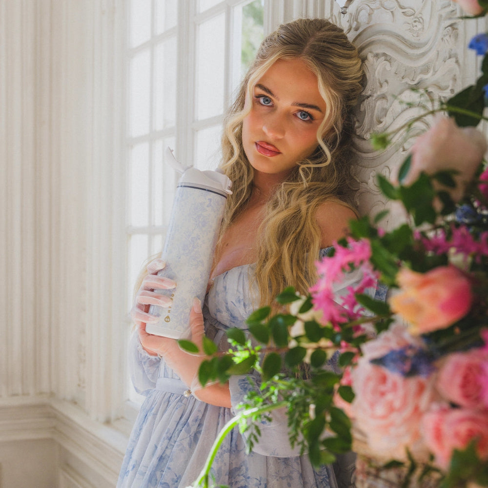 Woman holding a floral arrangement indoors with decorative wall and window in the background.