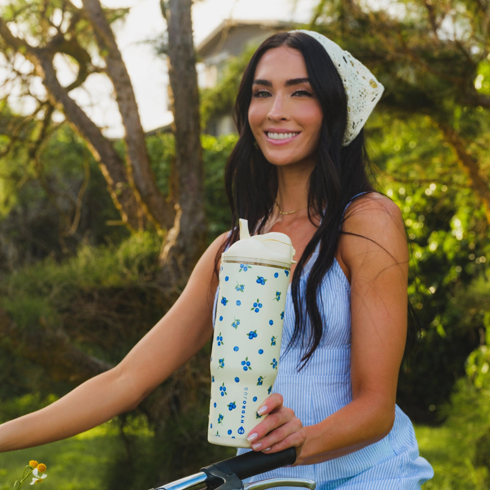 A woman outdoors on a bicycle holding a reusable insulated bottle with a cream lid and blue floral pattern. She is wearing a light blue striped top and a white patterned headscarf, with a blurred green garden background.