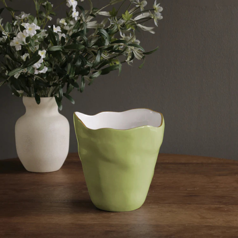 Green ceramic vase on a wooden surface with a white vase and flowers in the background.