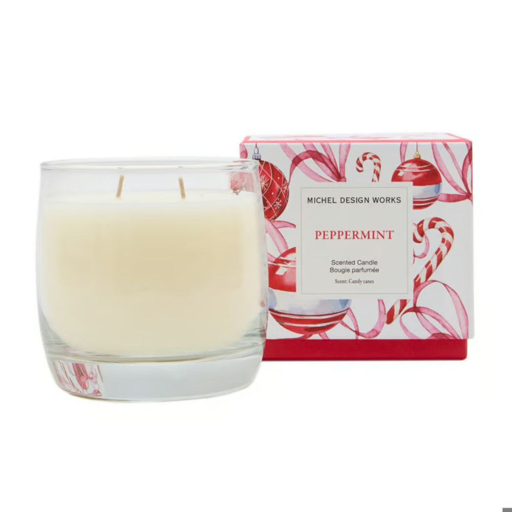 A Michel Design Works scented candle in a clear glass jar with two wicks, shown next to its red and white illustrated packaging box labeled Peppermint.
