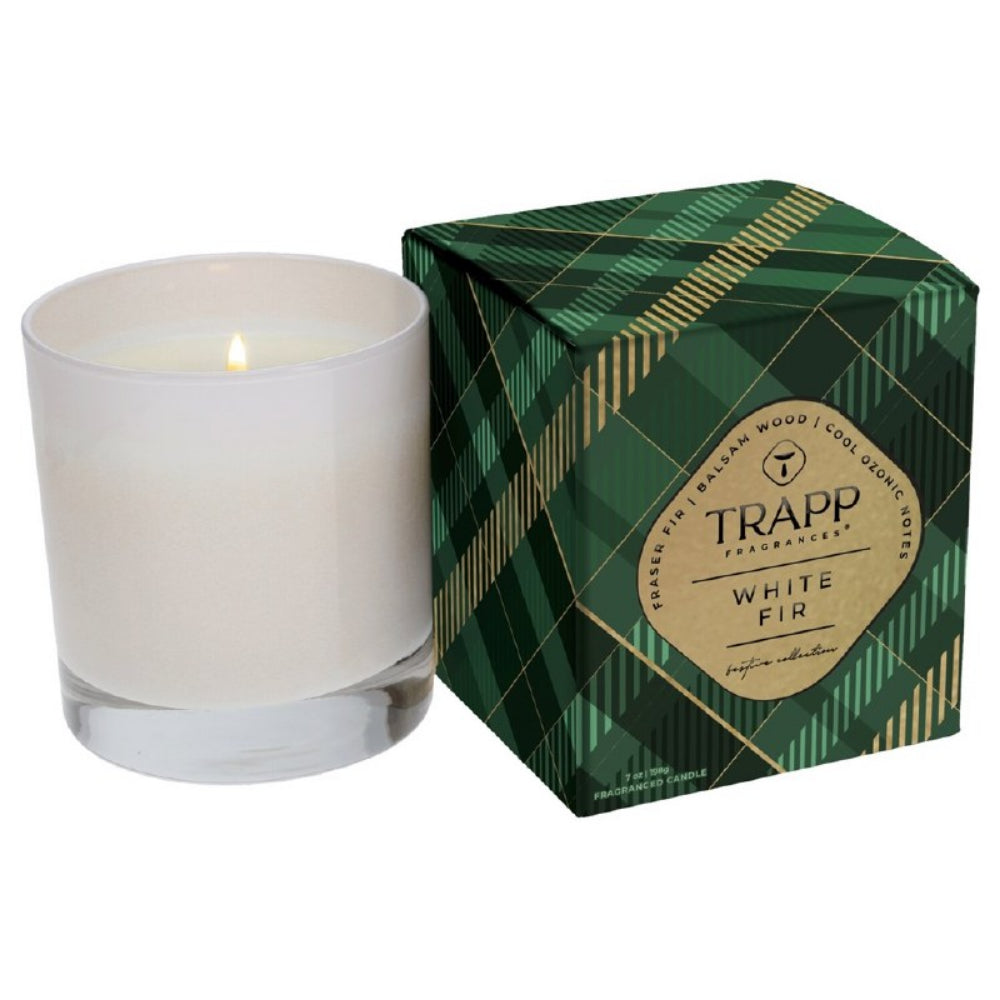 A lit white candle in a clear glass vessel next to its green plaid box with gold lines and a gold TRAPP fragrance label.