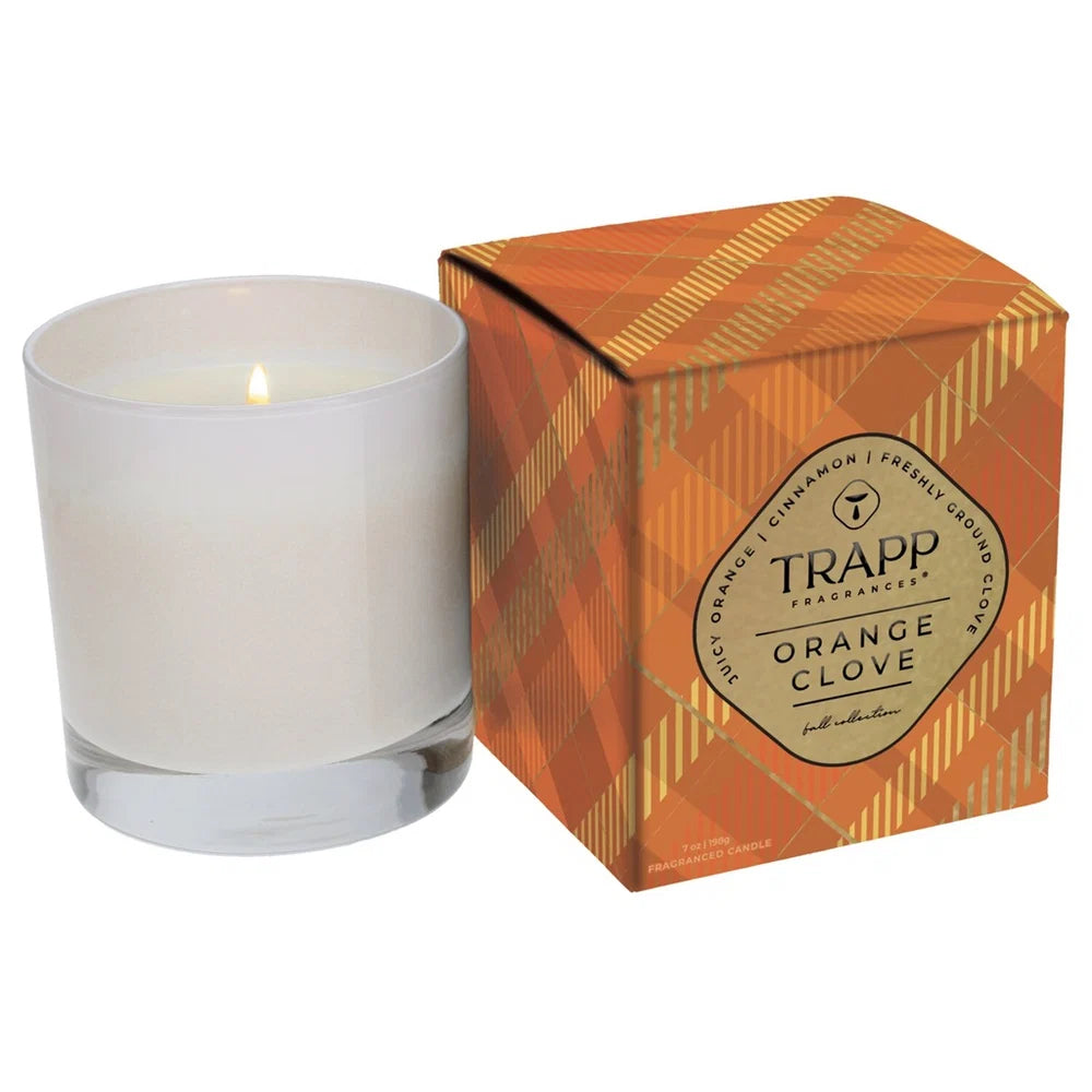 A lit white wax candle in a clear glass vessel next to its orange and gold patterned packaging box, which features the brand name TRAPP Fragrances and the scent Orange Clove.
