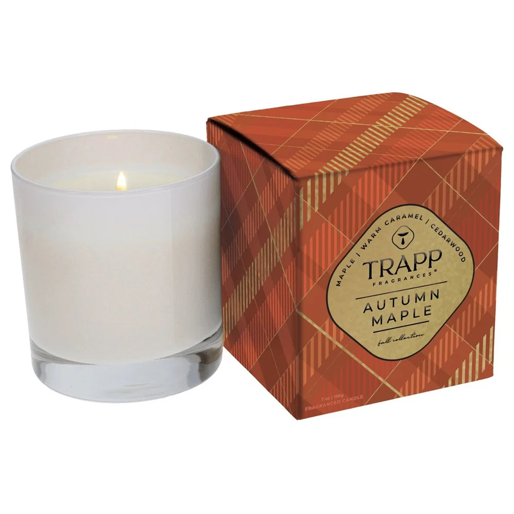 A lit white wax candle in a clear glass vessel next to its orange and gold patterned TRAPP Fragrances Autumn Maple box on a white background.