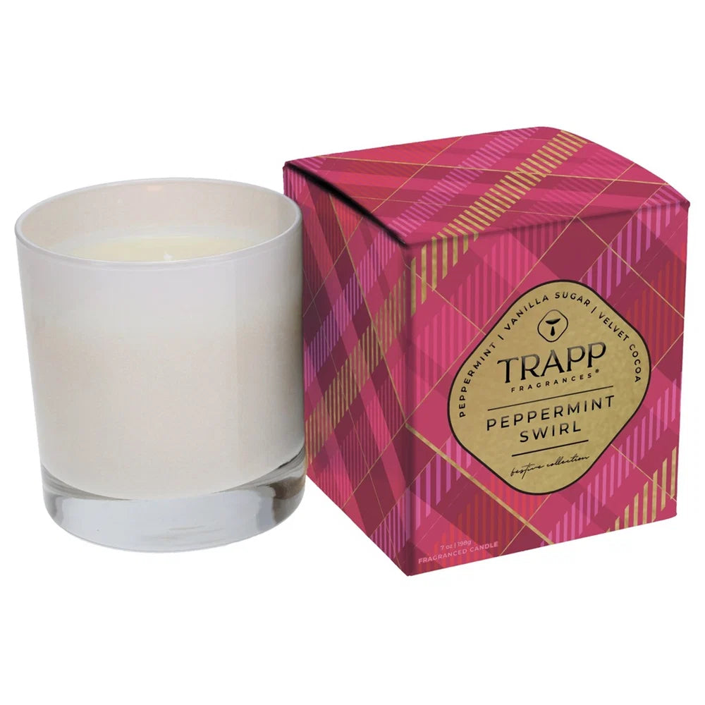 A white wax candle in a clear glass jar next to its pink and red plaid packaging box with a gold TRAPP Fragrances label.