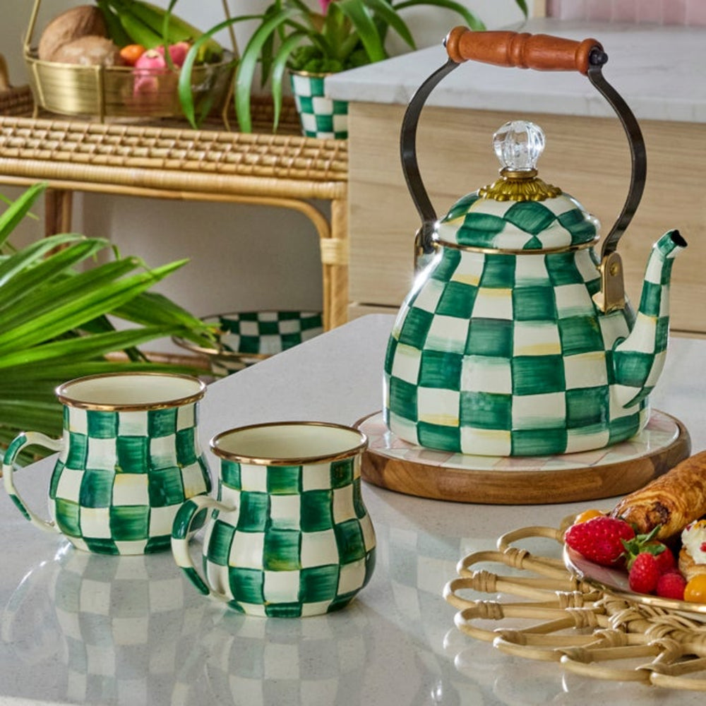 Green and white checkered teapot and mugs on a table with plants and food in the background