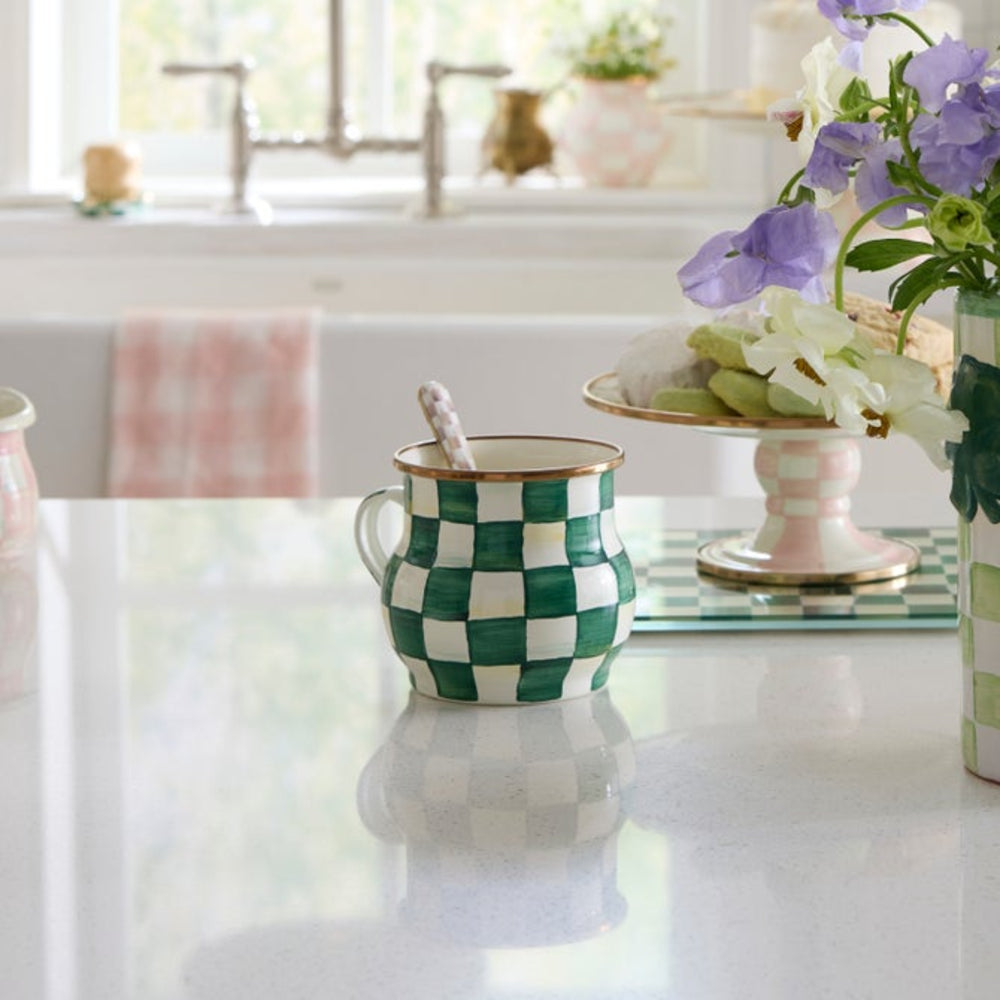 Green and white checkered mug on a kitchen counter with flowers and a cake stand in the background.