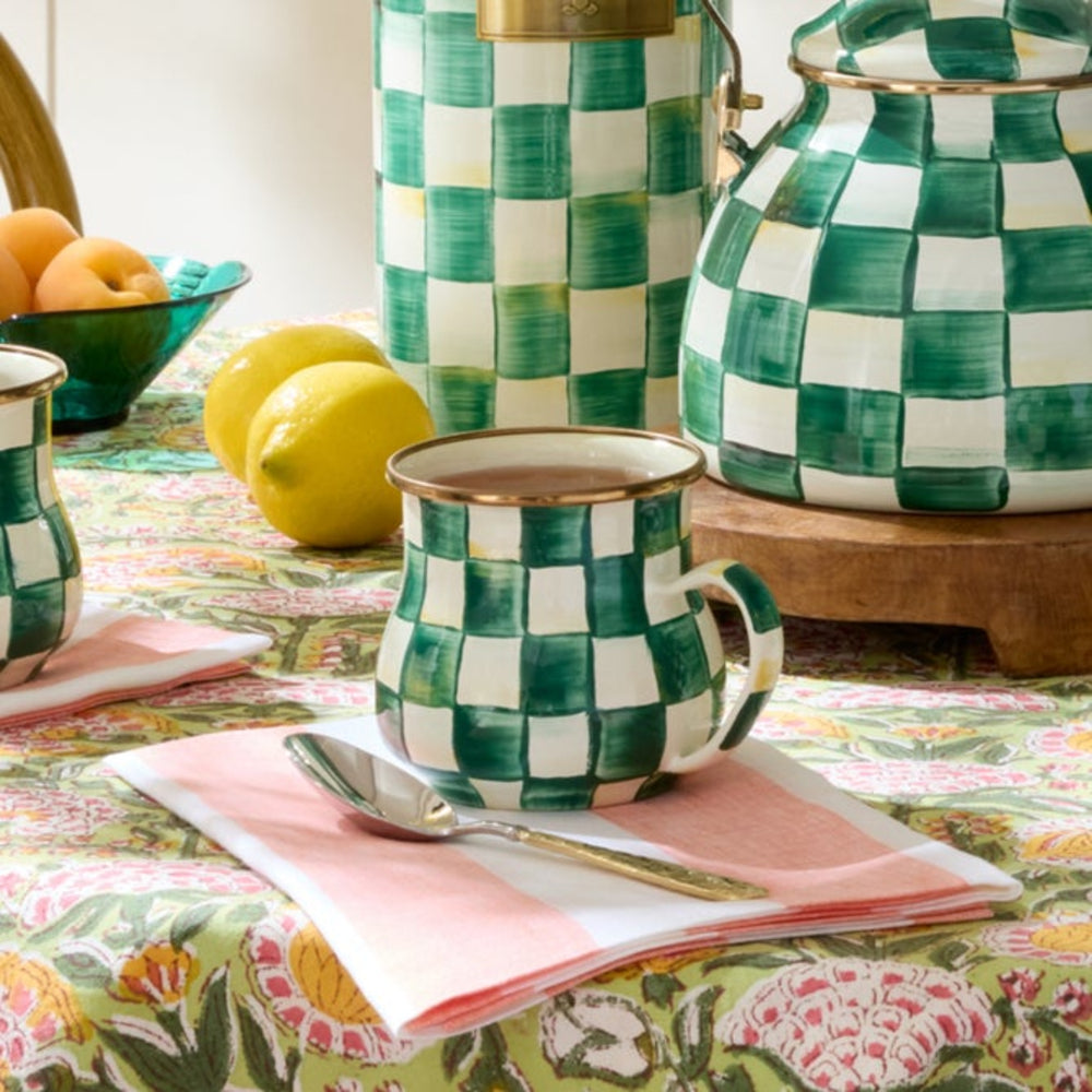 Green and white checkered mugs and teapot on a colorful tablecloth with lemons and a bowl of fruit.