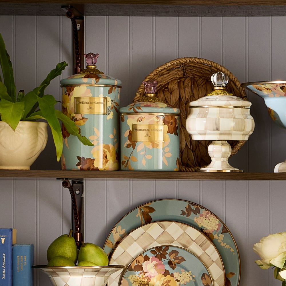 Decorative kitchen shelves with floral-patterned canisters, plates, and a plant against a gray wall.