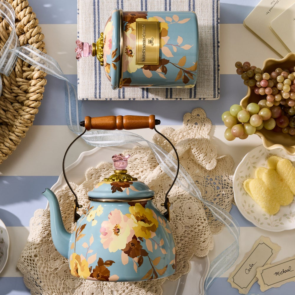 Floral-patterned teapot on a decorative table with a striped cloth and fruit.