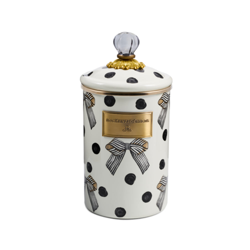 A white enamel canister with black polka dots, striped ribbon motifs, and a gold-tone front plaque, topped with a clear glass knob and gold trim, shown on a white background.