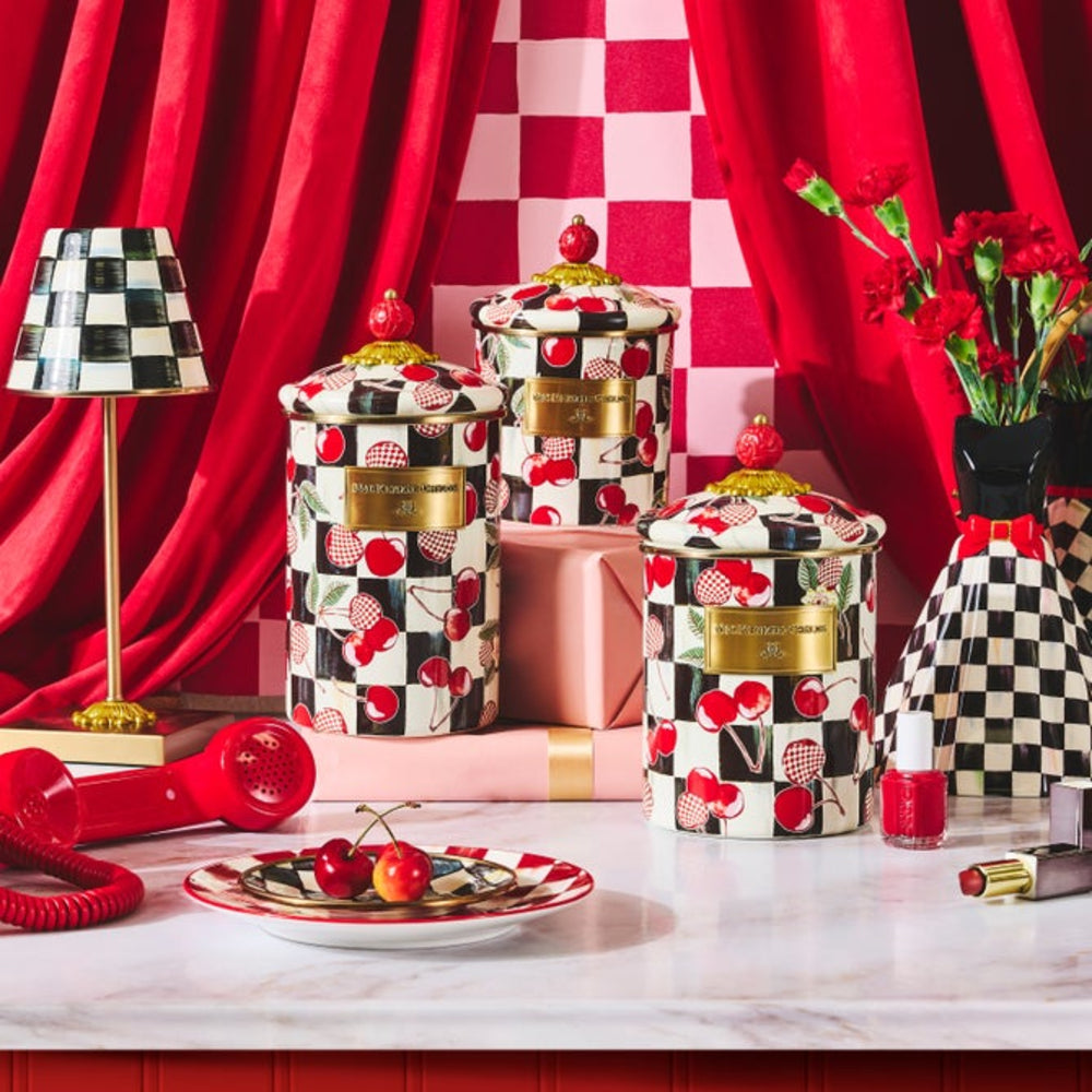 Three decorative cylindrical jars with lids, covered in a black-and-white checkerboard and cherry pattern, arranged on a white marble surface with red and pink props, including a red hair dryer, flowers, and a checkered lamp.