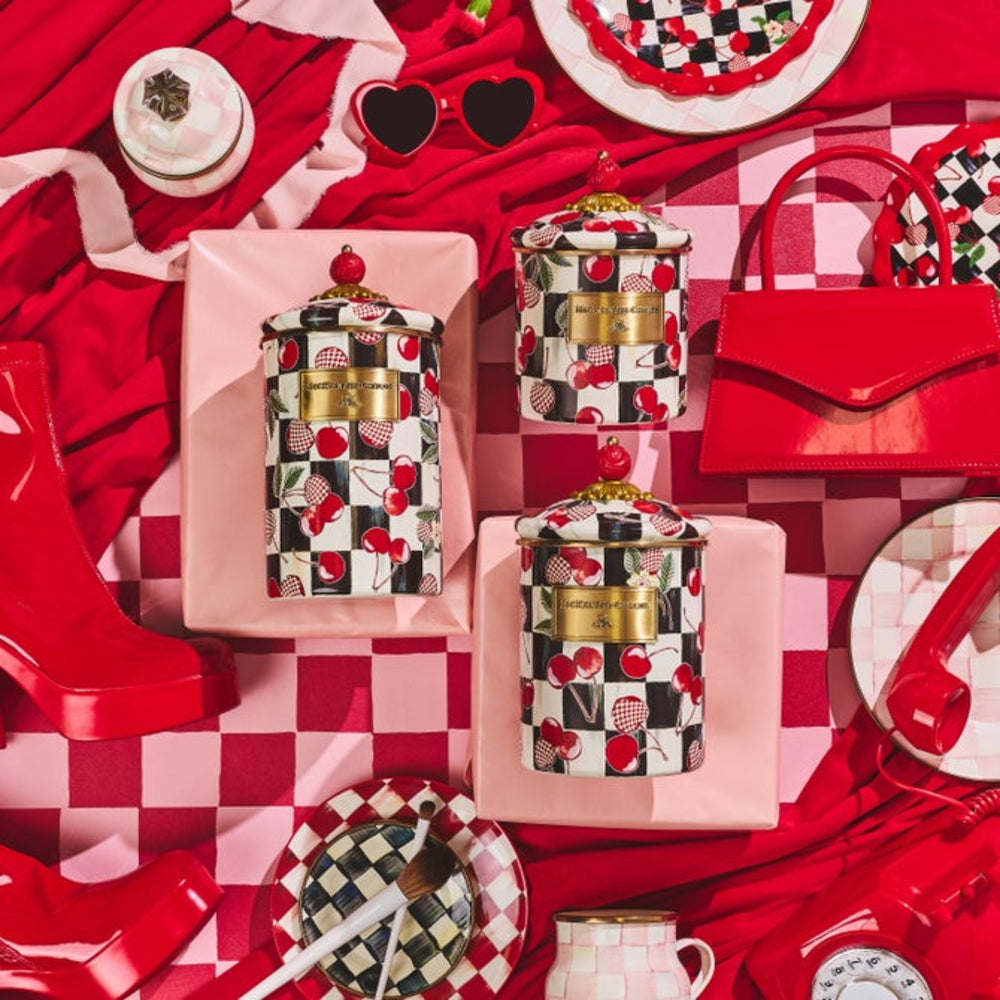 A styled flat-lay featuring three patterned ceramic canisters with gold lids, surrounded by red and pink accessories including a red handbag, red boots, a red-and-white checkered cloth, and red heart sunglasses on a red background.