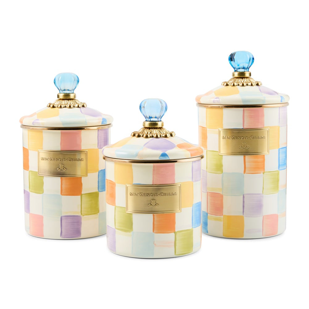 Three ceramic canisters with lids in a pastel multicolor checkerboard pattern, featuring gold-tone metal plaques and blue glass knob lids, set against a plain white background.
