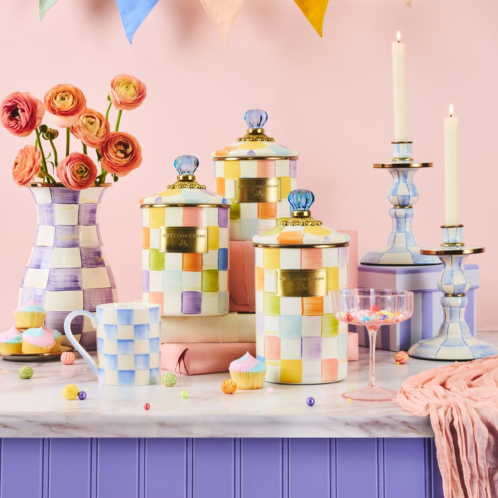 A styled tabletop scene with pastel checkered ceramic canisters, a matching mug, flowers in a vase, candlesticks, and cupcake decorations against a pink wall and lavender cabinet.
