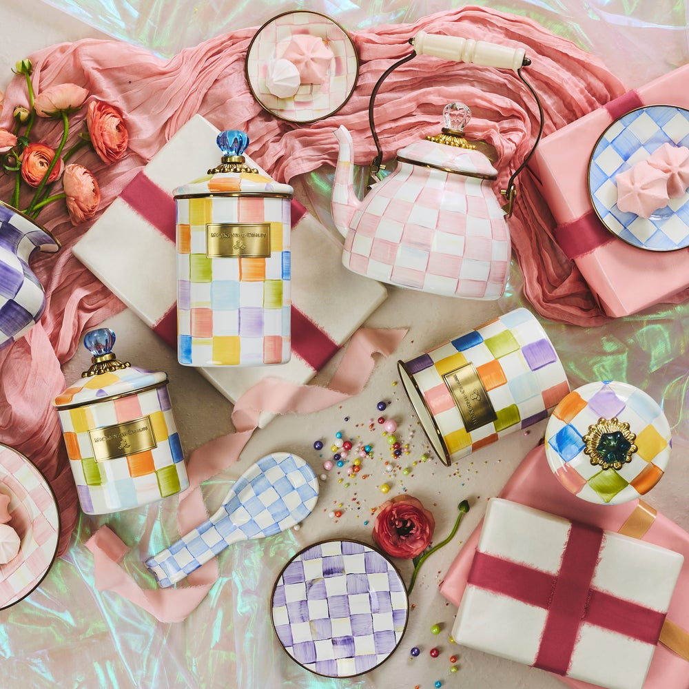 A styled flat-lay of pastel pink and multicolor checkered ceramic tea and coffee accessories, including a teapot, mugs, cups, spoons, and boxes, arranged with pink fabric, flowers, and confetti on a shiny iridescent surface.