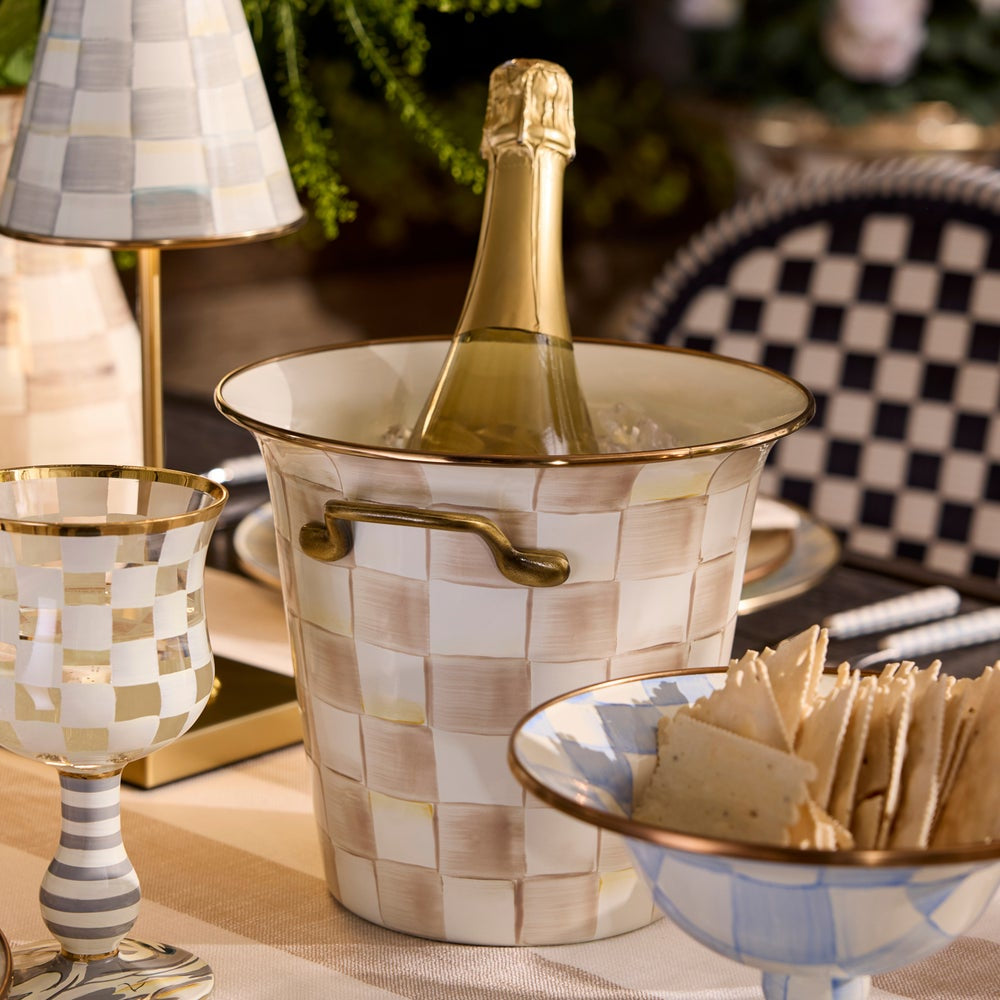 Bottle of champagne in a decorative ice bucket on a table with crackers and a lamp.