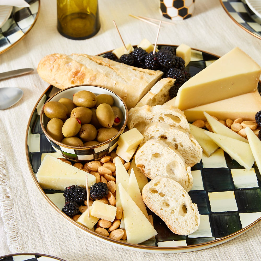 Assorted cheese, bread, olives, and nuts on a checkered plate with a bottle of wine in the background.