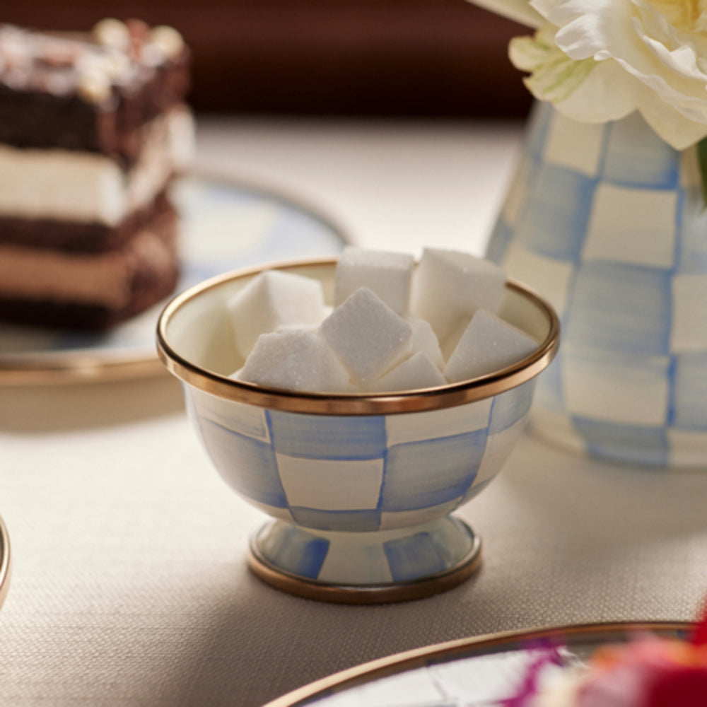 A small footed bowl with a blue and white checkered pattern and gold trim, filled with white sugar cubes, set on a table with a blurred cake and vase in the background.
