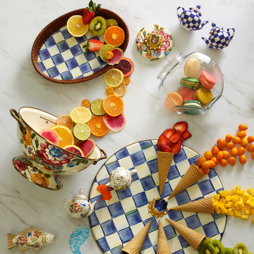 MacKenzie-Childs royal check rattan & enamel tray on a table with a colorful arrangement of fruits, cookies, and decorative items on a  on a marble surface.