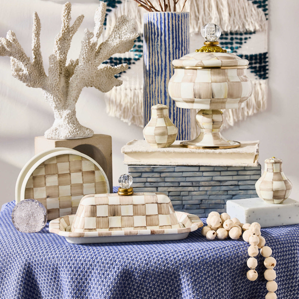 A beige and brown Mackenzie-Childs Mocha Check Napkin Holder made of enameled steel with a hand-painted, coffee-colored check pattern on a blue table cloth with other simular items on it.