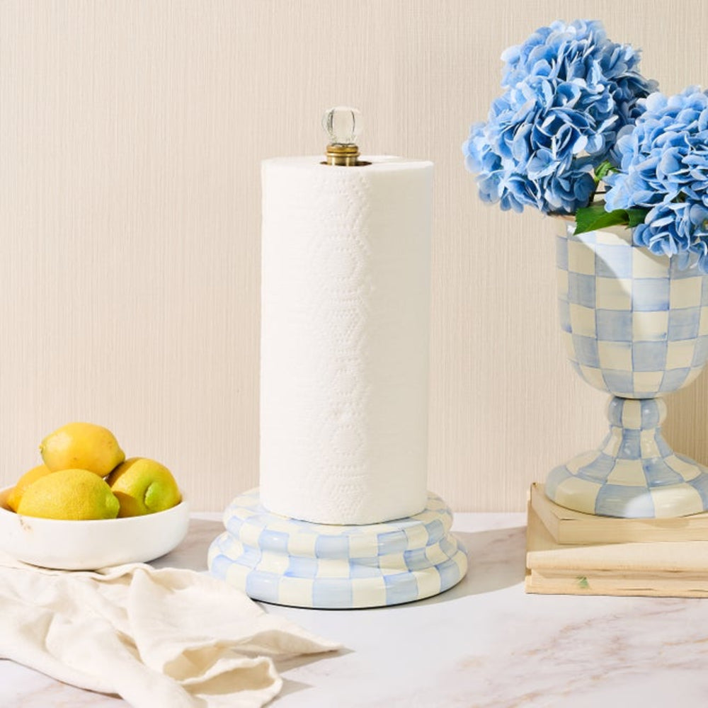 A paper towel holder with a roll of white paper towels, styled on a marble-like surface with a bowl of lemons, a folded cream cloth, and a blue-and-white checkered vase of flowers in the background.