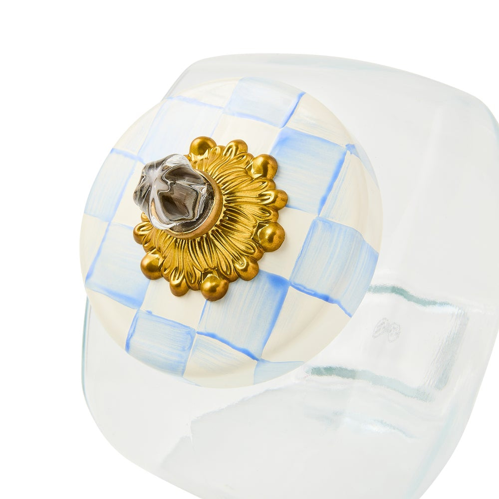 A close-up of a clear glass ring featuring a circular insert with a light blue and white checkered pattern, adorned with a prominent textured gold sunburst medallion and a central silver-toned emblem.
