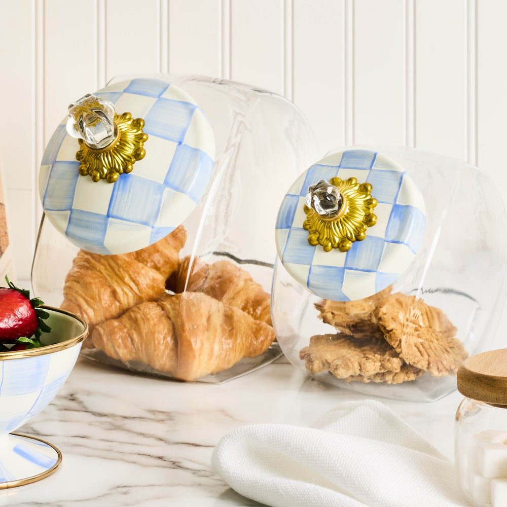 Two clear glass food storage canisters with round lids decorated in a light blue and white checkered pattern and topped with ornate gold-tone knobs, shown on a marble countertop with pastries and a strawberry inside.