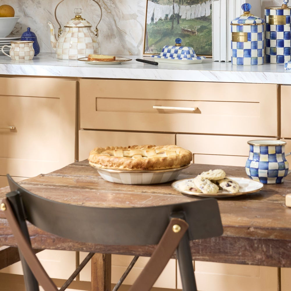A rustic wooden dining table in a warm beige kitchen setting, featuring a baked apple pie in a metal dish and a small plate of cookies. A dark brown leather and wood chair is in the foreground, with decorative ceramic canisters and a teapot on the marble countertop behind.