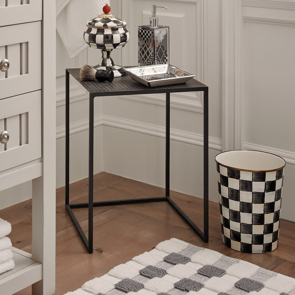 Bathroom setting with a small table, checkered trash can, and decorative items.
