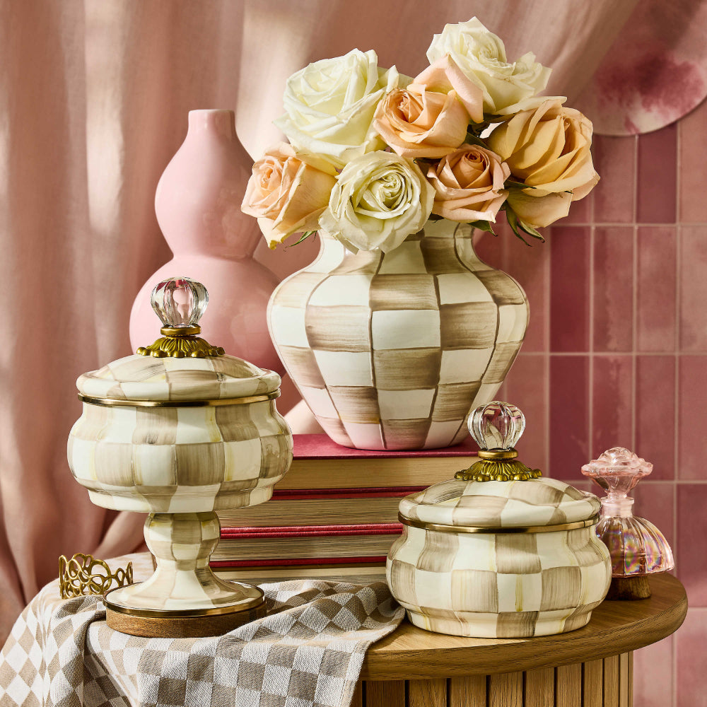 Mackenzie-Childs Mocha Check Curiosity Pot with clear top handle on a table with other mocha check enamelware with red books in a decorative pink setting. 