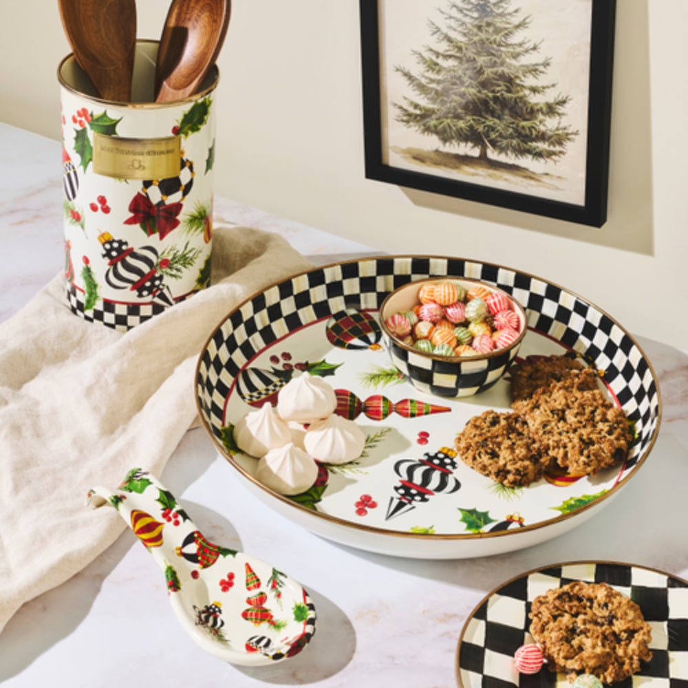 A styled tabletop scene with a large oval holiday-themed serving platter, a matching small plate, a spoon, and a utensil holder. The platter and holder have a white base with black-and-white checkered trim, red berries, green holly, plaid accents, and festive illustrations.