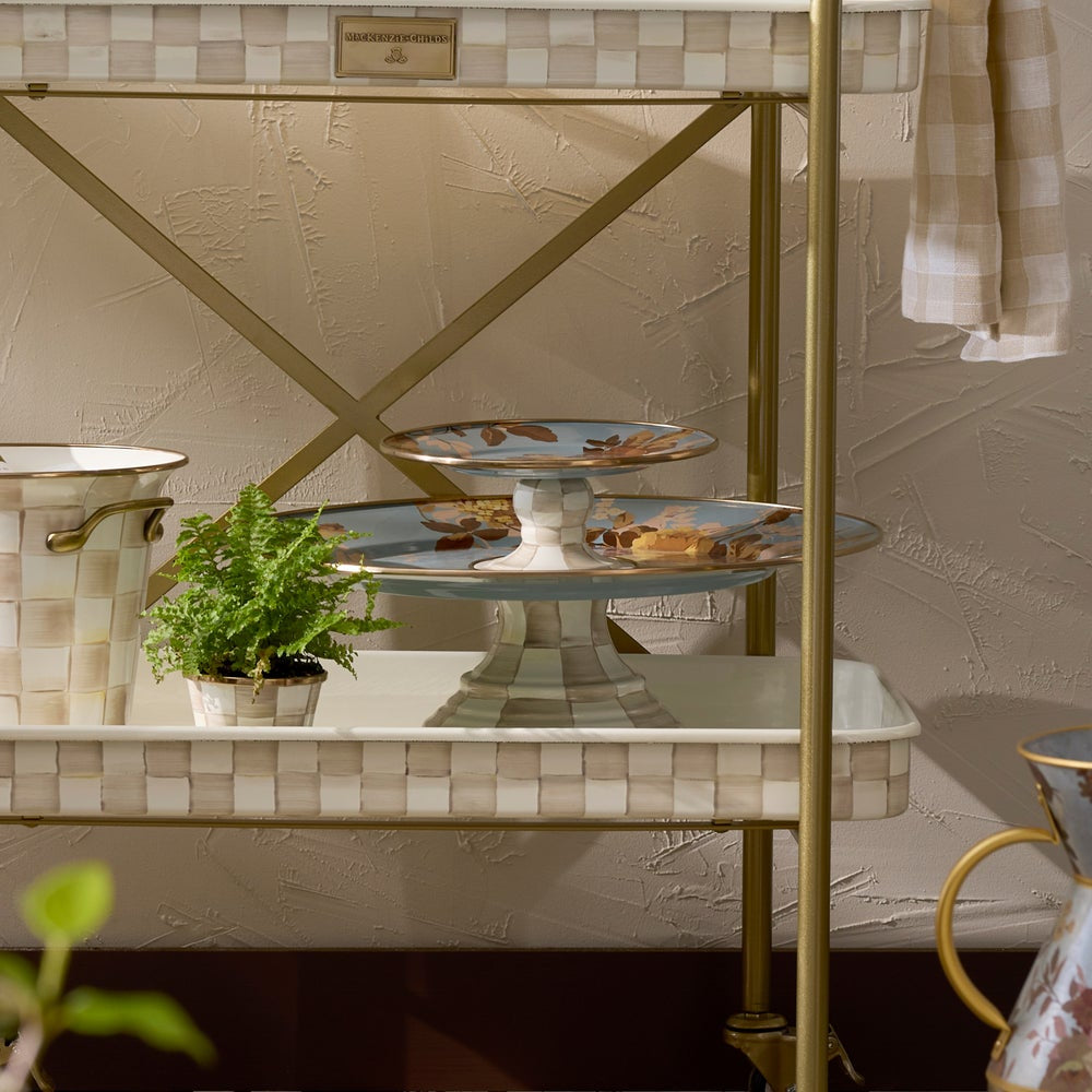 A brass and cream checkered two-tier serving cart with a white marble-look shelf, styled with a small potted green plant, a decorative cake stand, and a patterned ceramic pitcher against a textured beige wall.