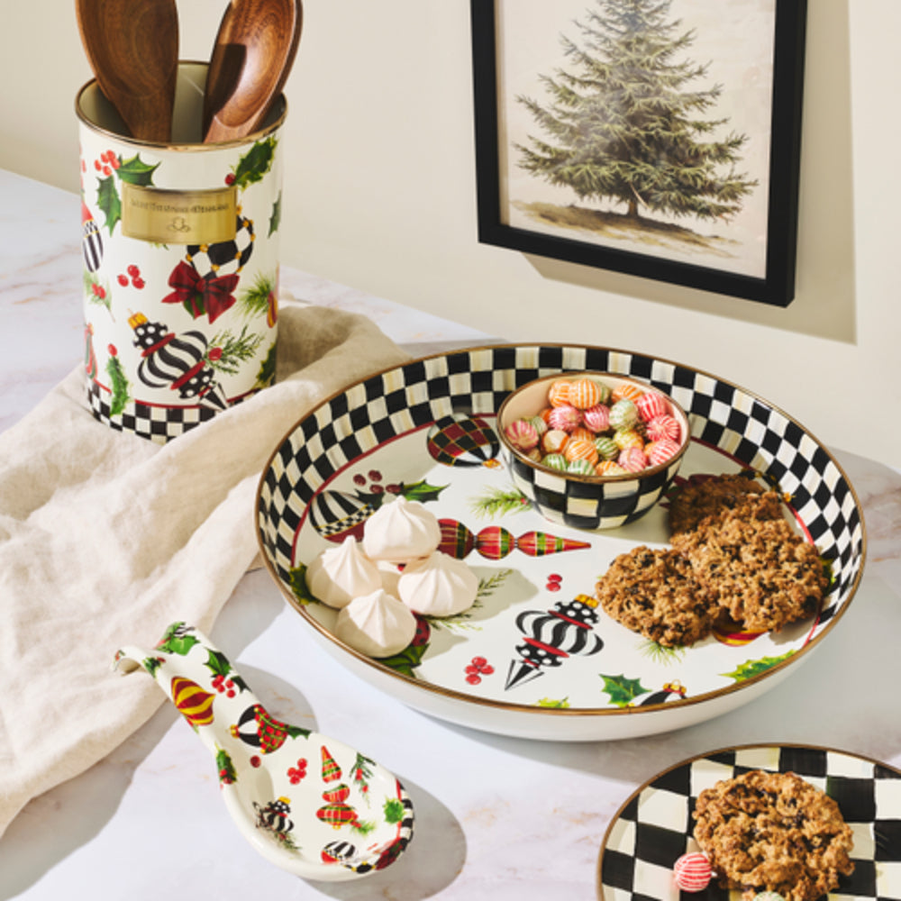 A styled tabletop scene showing a decorative ceramic serving set: a large round platter with a black-and-white check border, a matching small bowl, a spoon, and a utensil holder with wooden spoons. The ceramic pieces are white with holiday motifs including holly, pinecones, acorns, and red berries.