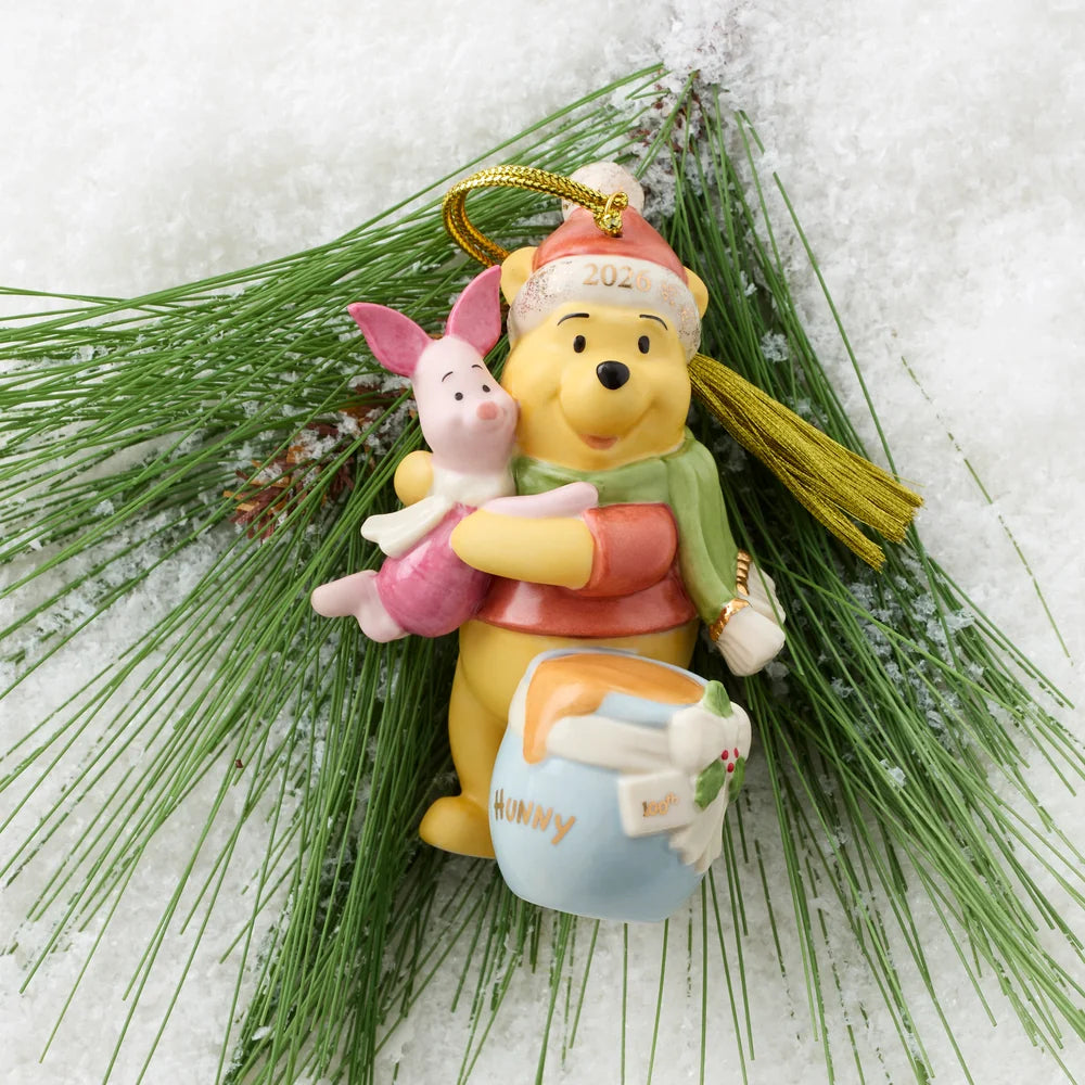 A Winnie the Pooh bear Christmas ornament holding Piglet and a honey pot, styled on green pine branches with white snow-like background.