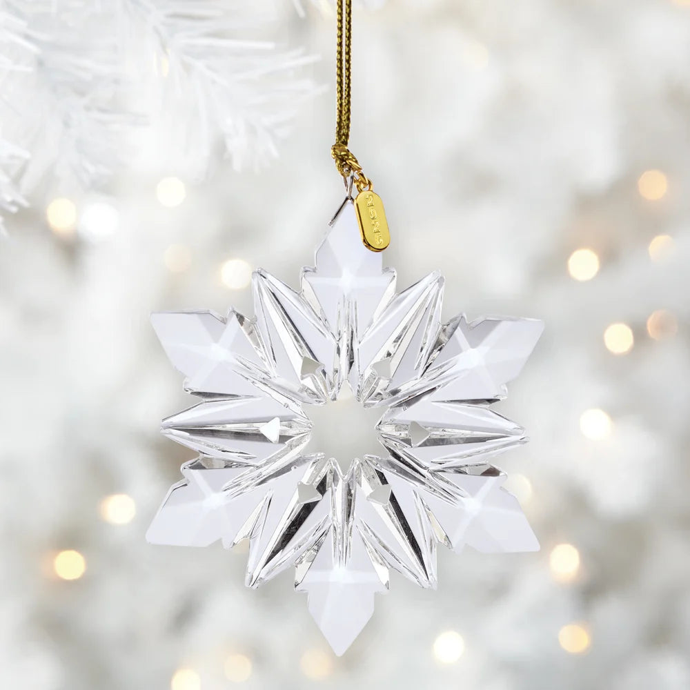 A clear faceted snowflake Christmas ornament hanging from a gold string with a small gold tag, set against a soft white blurred Christmas tree background with warm bokeh lights.